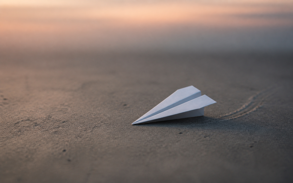 A paper plane resting on sand at dusk with its flight path trailing behind it